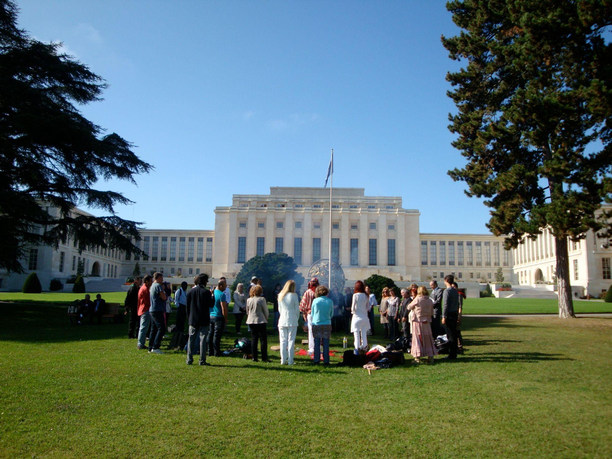 Eine Gruppe steht im Kreis um ein Ritualfeuer auf dem Rasen vor dem Palais des Nations der UNO in Genf