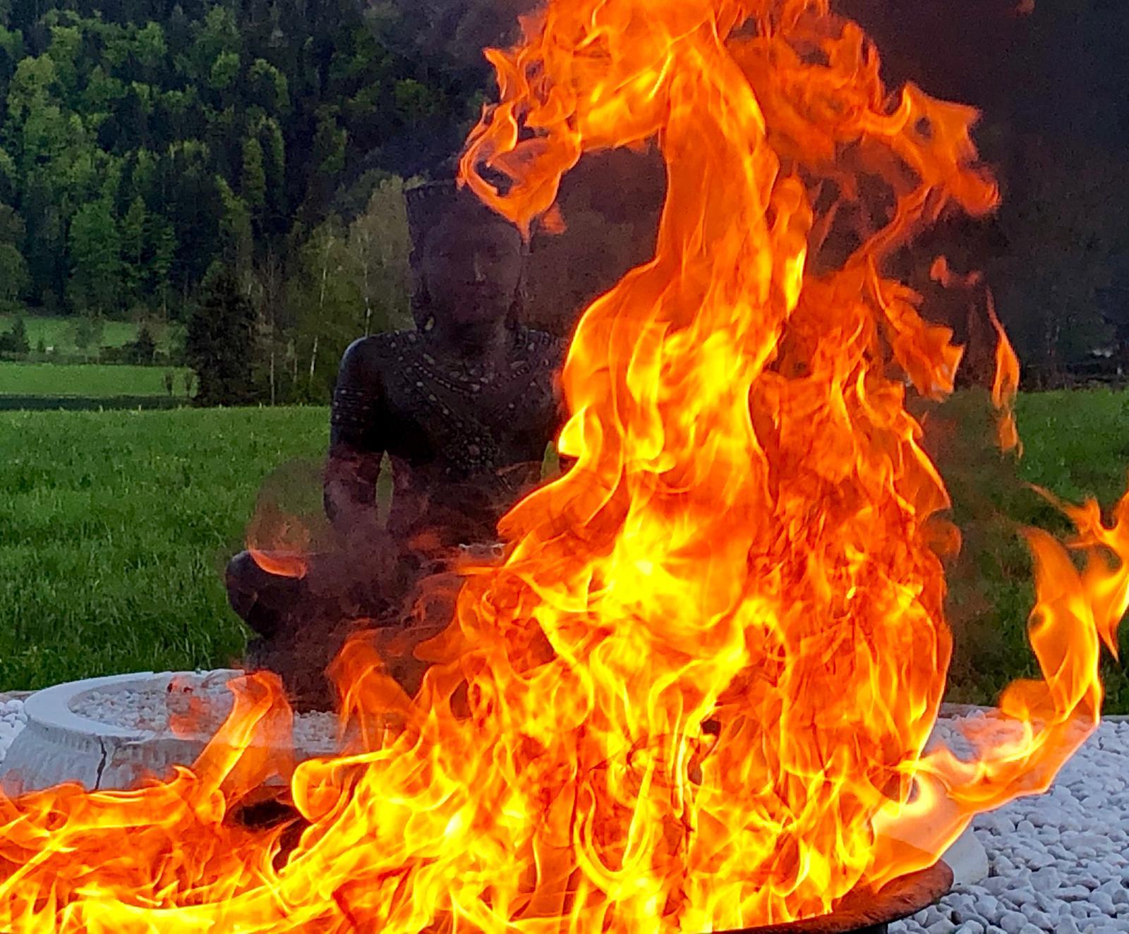 Hohe Flammen eines Ritualfeuers, dahinter eine dunkle Steinstatue im Lotossitz vor grünen Wiesen und Wald