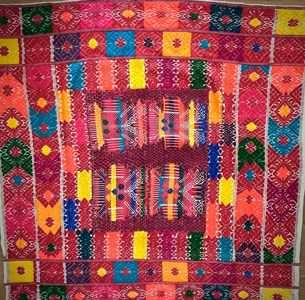 Eine handgewebte Decke aus dem Webeprojekt, quadratisch, mit leuchtenden Rauten und Bändern in Rot, Gelb, Türkis, Grün und Orange