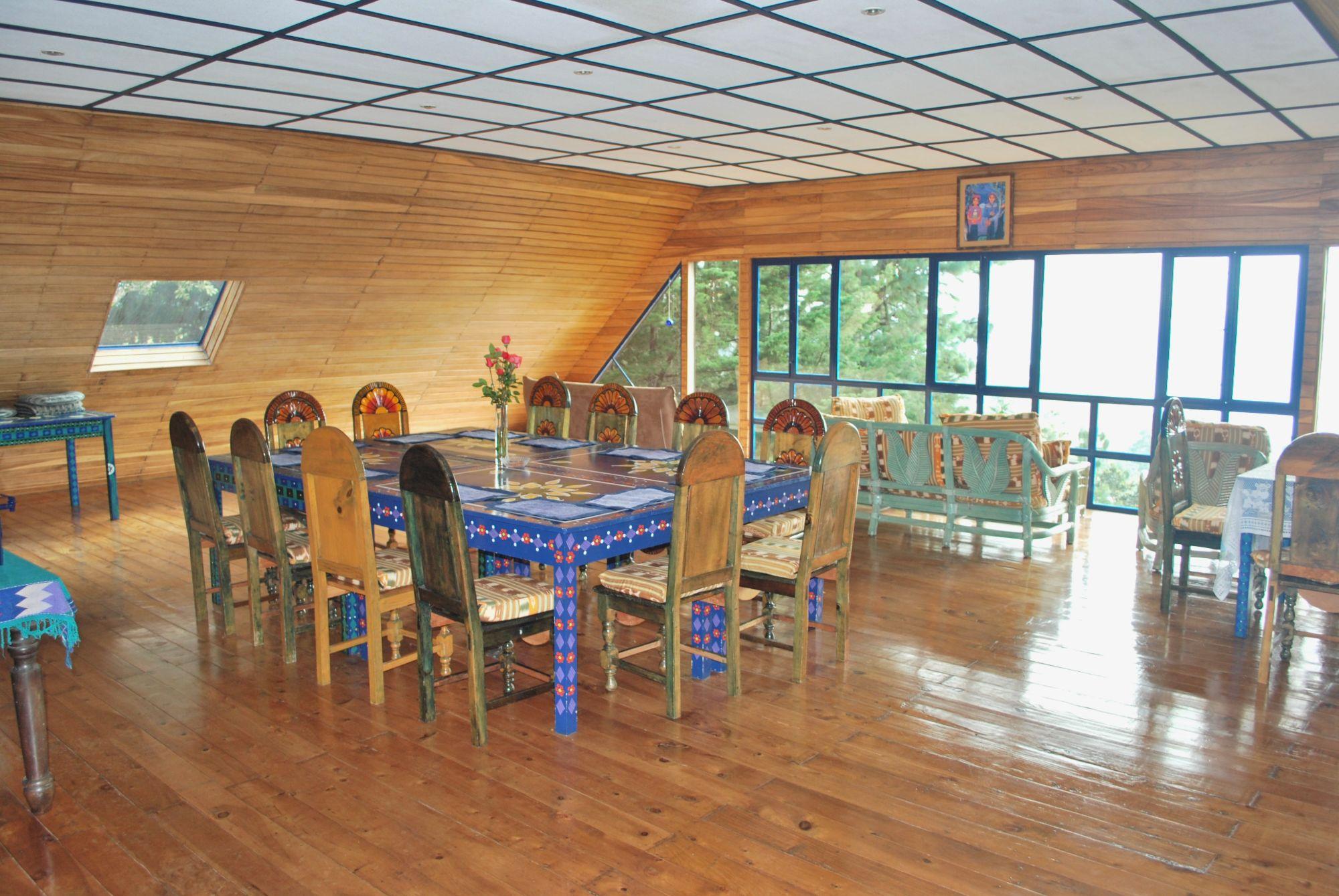 Die Casa Kawoq: ein heller Holzraum mit großen Fenstern ins Grüne, einem bemalten blauen Tisch mit geschnitzten Stühlen und Sofas
