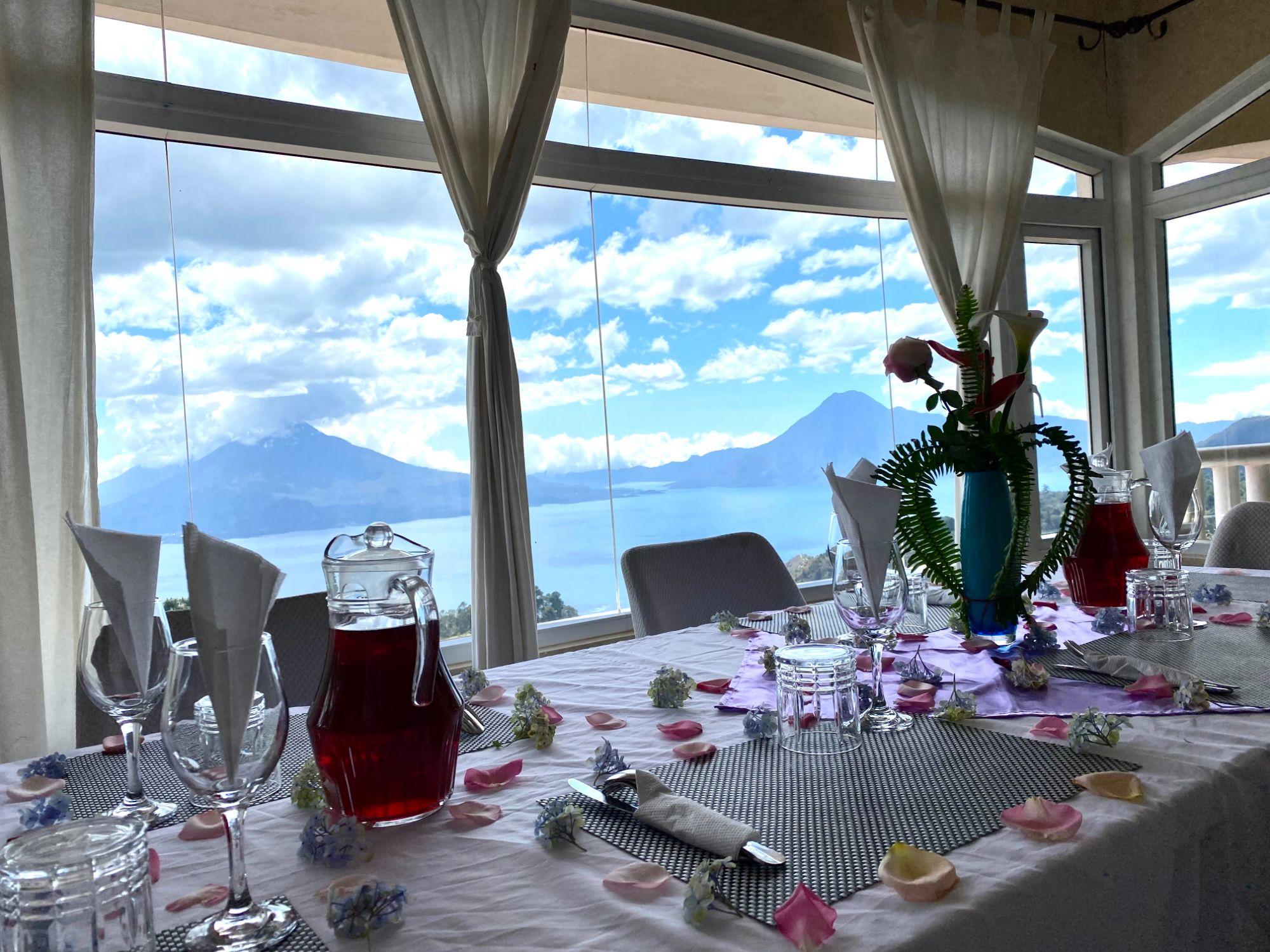 Ein gedeckter Tisch mit Blütenblättern im Speisesaal, durch die großen Fenster der Atitlánsee und die Vulkane