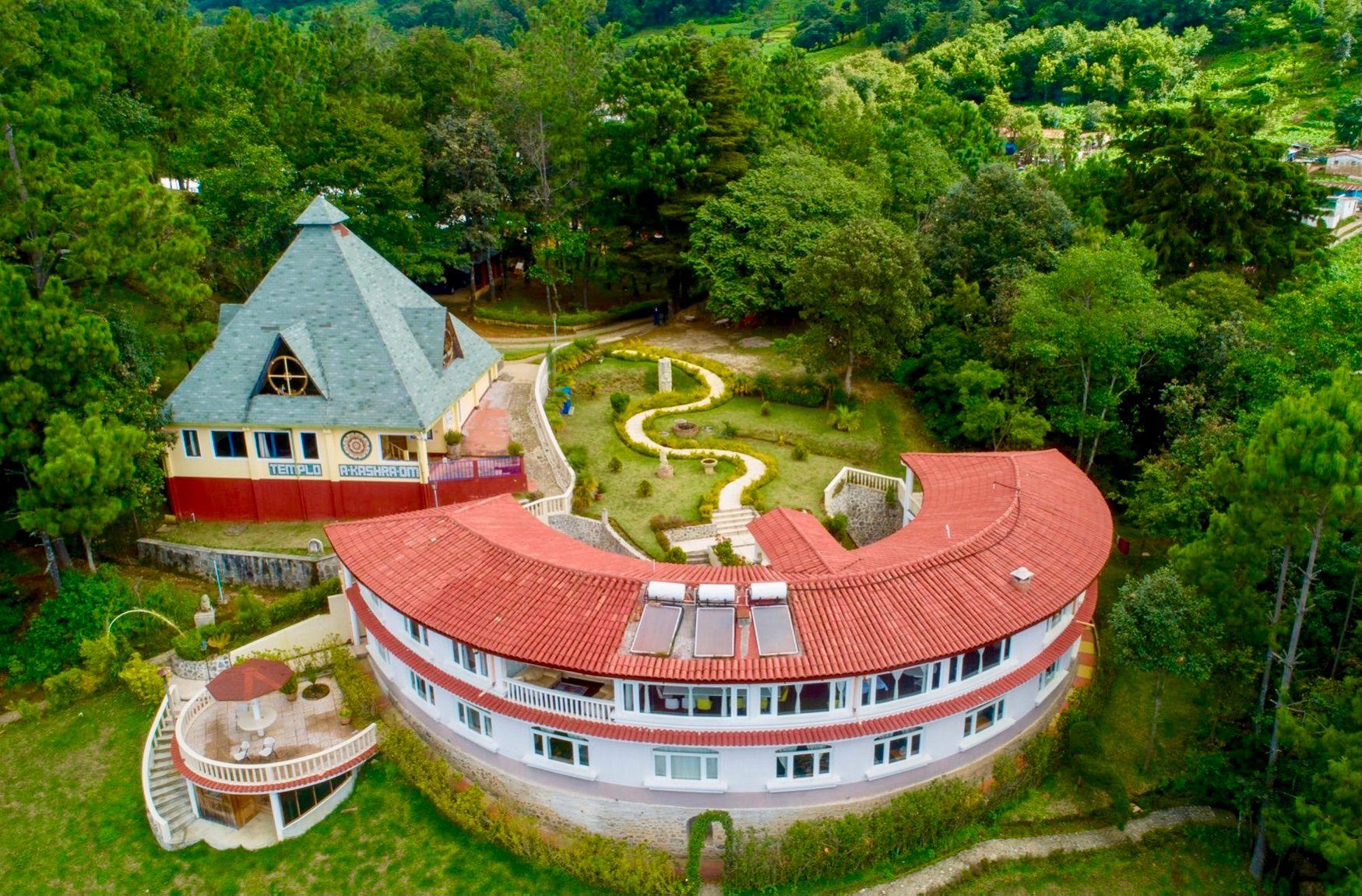 Das Zentrum PAZ MUNDO von oben: der geschwungene Hauptbau mit rotem Ziegeldach, der Tempel A-KASHRA-OM mit grünem Pyramidendach und der Garten mit dem Schlangenweg, ringsum Wald